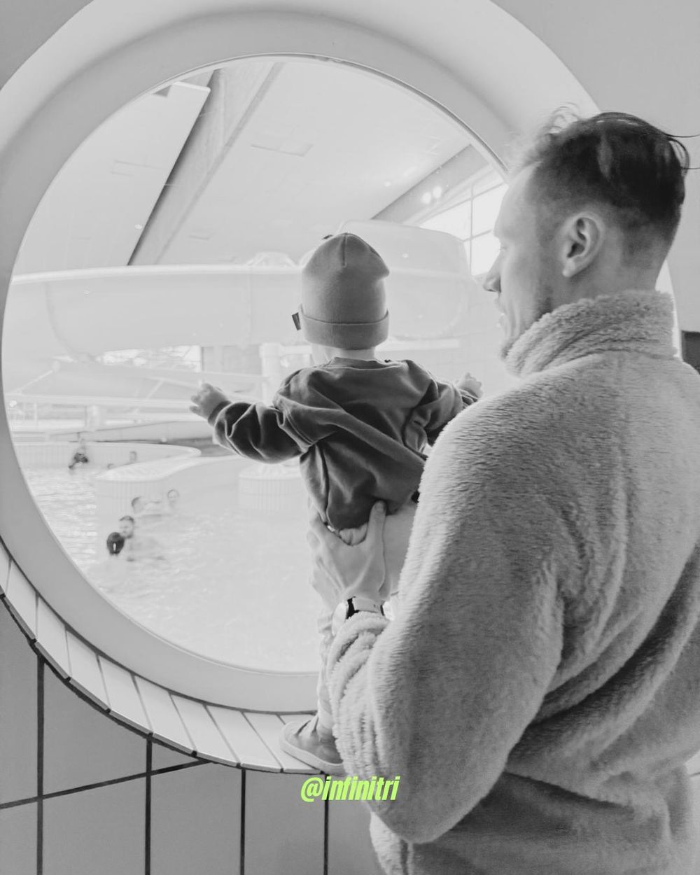Un membre du club porte son enfant devant le hublot d'une piscine, tous deux regardent les nageurs